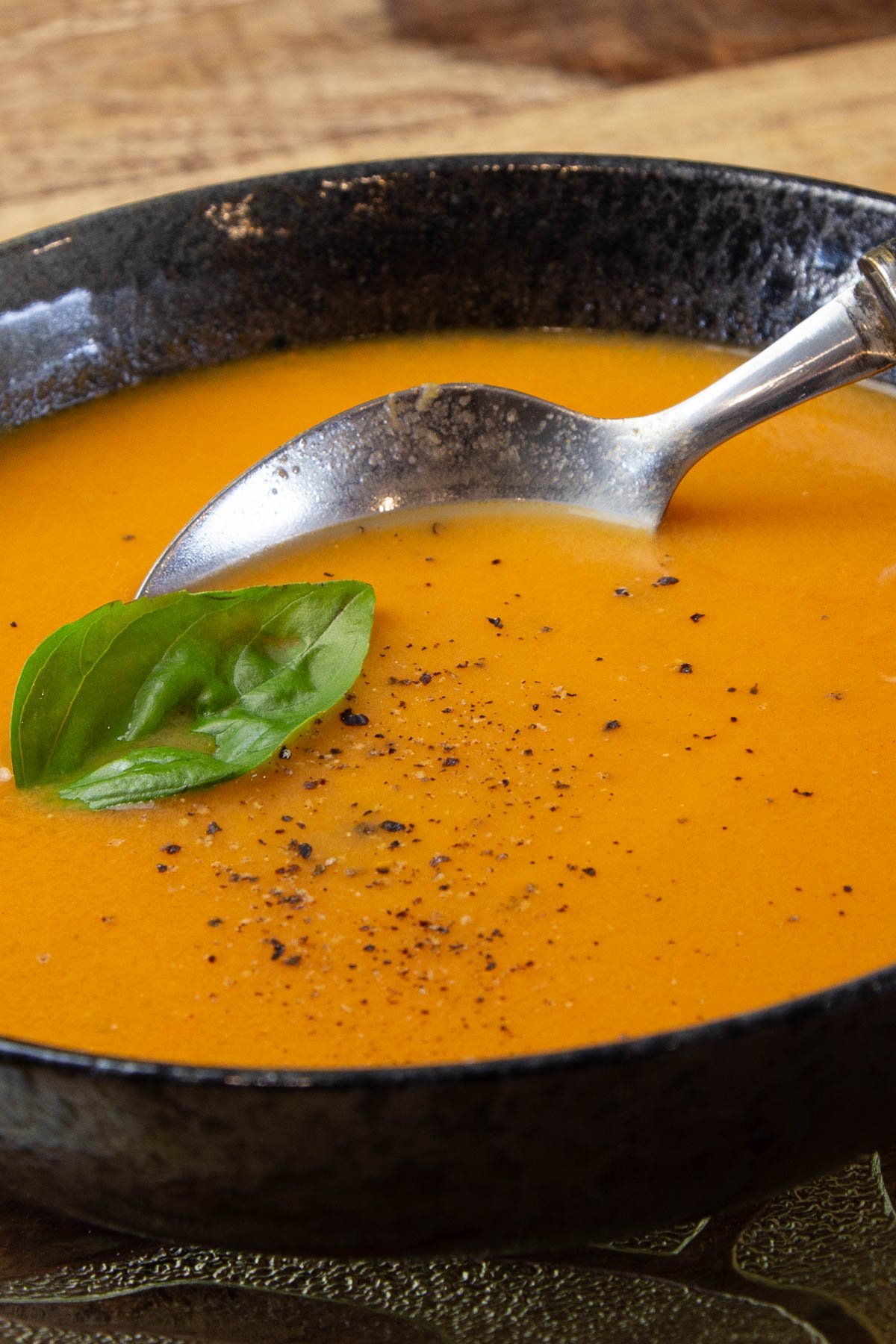 A black bowl filled with tomato soup, garnished with a fresh basil leaf. A silver spoon dips into the soup, with cracked pepper sprinkled on top.