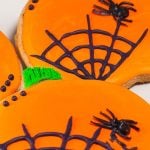 Pumpkin-shaped cookies decorated with bright orange icing, black spiderweb designs, and small black spiders for a festive Halloween treat.