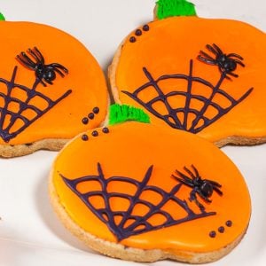 Pumpkin-shaped cookies decorated with bright orange icing, black spiderweb designs, and small black spiders for a festive Halloween treat.