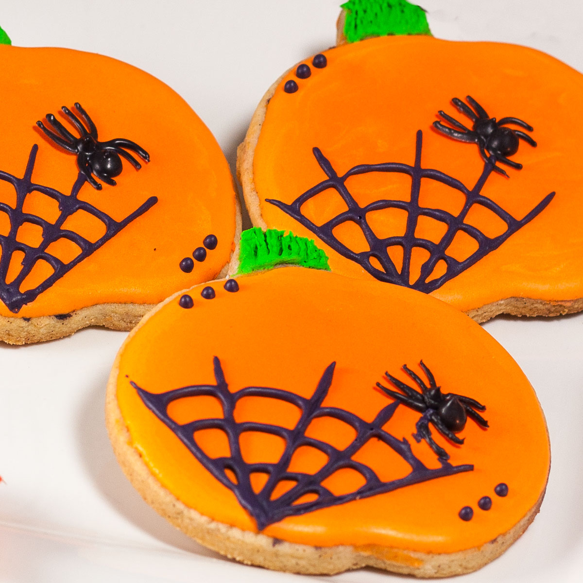 Pumpkin-shaped cookies decorated with bright orange icing, black spiderweb designs, and small black spiders for a festive Halloween treat.