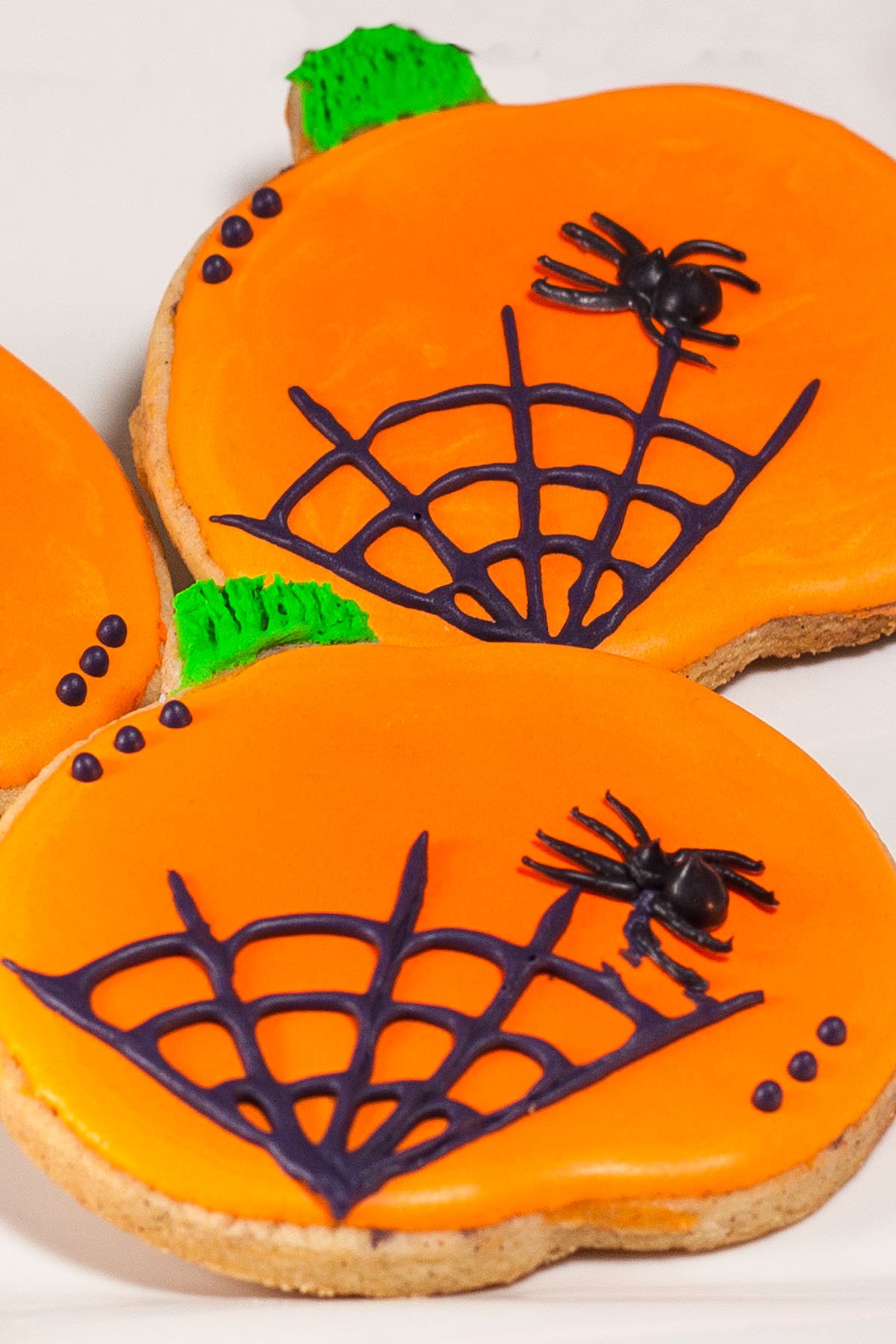 Pumpkin-shaped cookies decorated with bright orange icing, black spiderweb designs, and small black spiders for a festive Halloween treat.