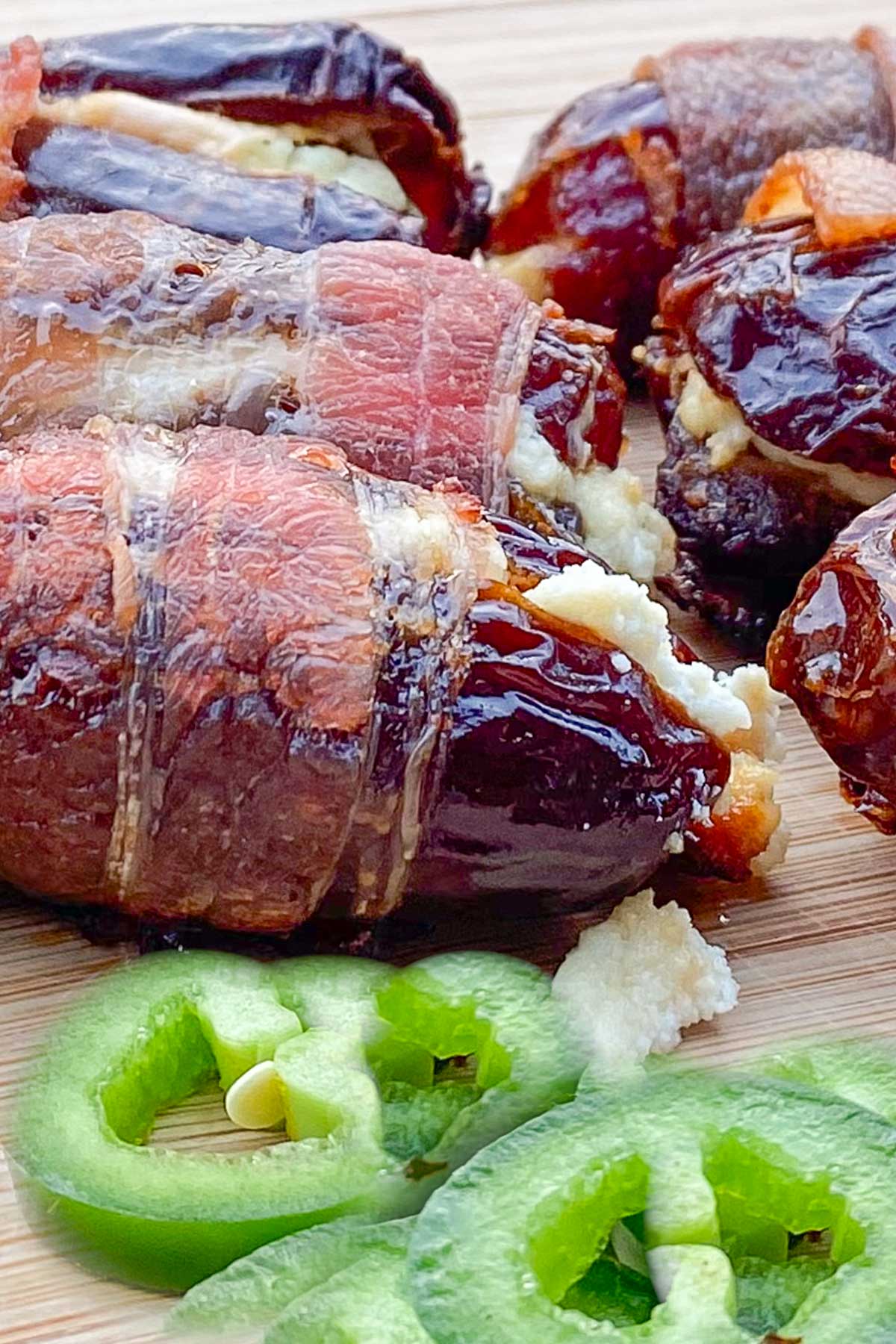 Crisp bacon-wrapped dates stuffed with cheese are placed on a wooden board. Sliced jalapeños are in front.