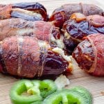 Crisp bacon-wrapped dates stuffed with cheese are placed on a wooden board. Sliced jalapeños are in front.