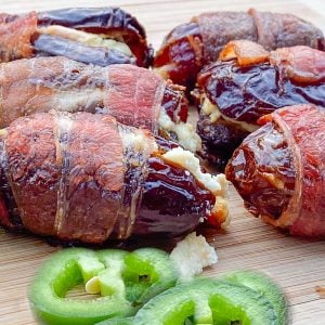 Crisp bacon-wrapped dates stuffed with cheese are placed on a wooden board. Sliced jalapeños are in front.