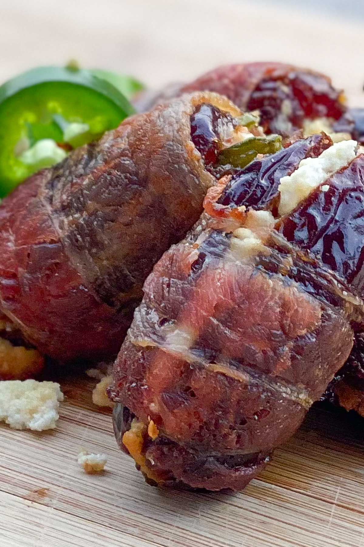 Crisp bacon-wrapped dates stuffed with cheese are placed on a wooden board. Sliced jalapeños along side.