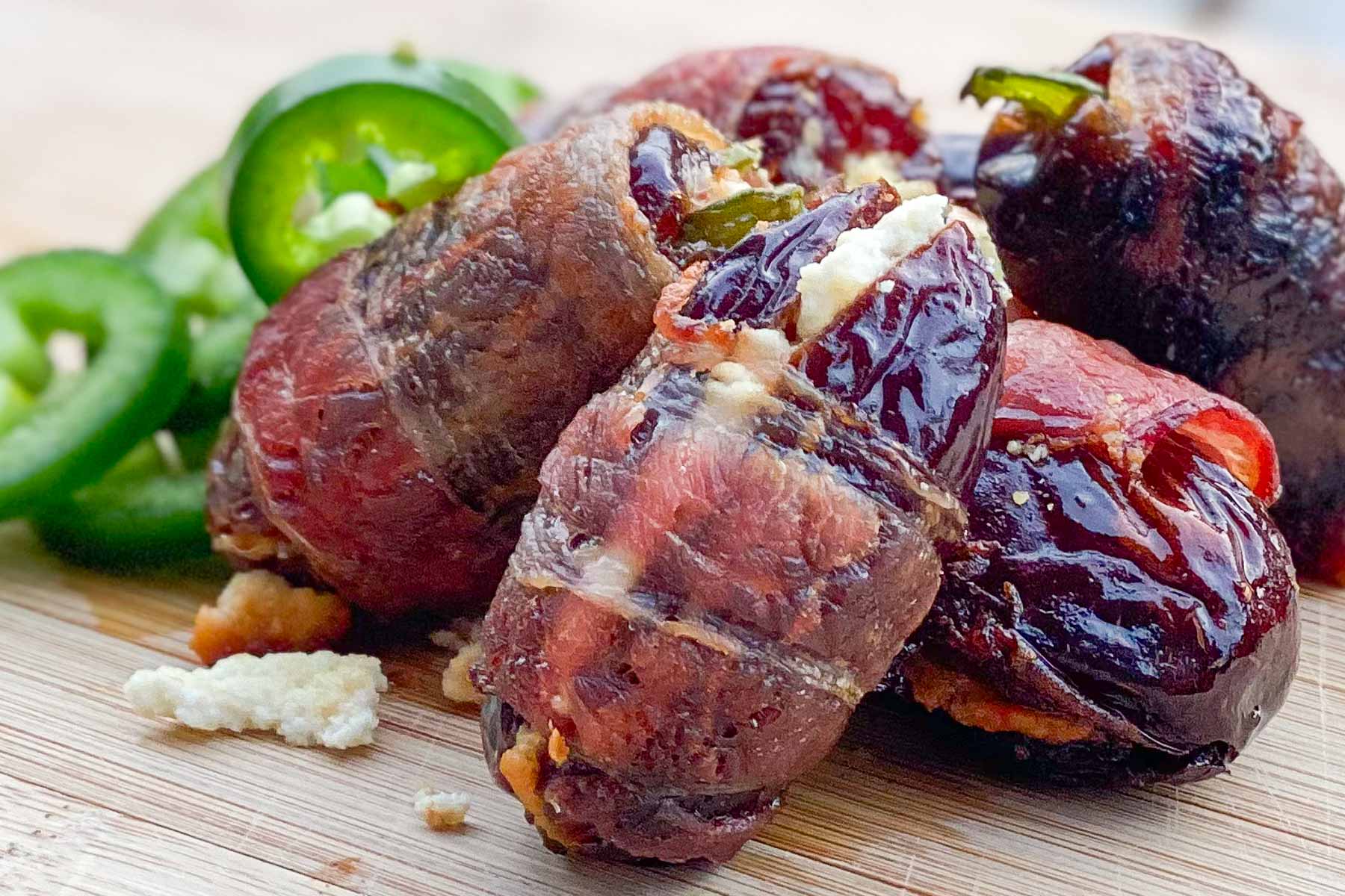 Crisp bacon-wrapped dates stuffed with cheese are placed on a wooden board. Sliced jalapeños along side.