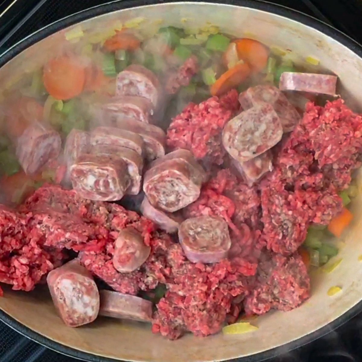 Raw ground meat and sliced sausage are sizzling in a dutch over with onions, carrots, and celery.