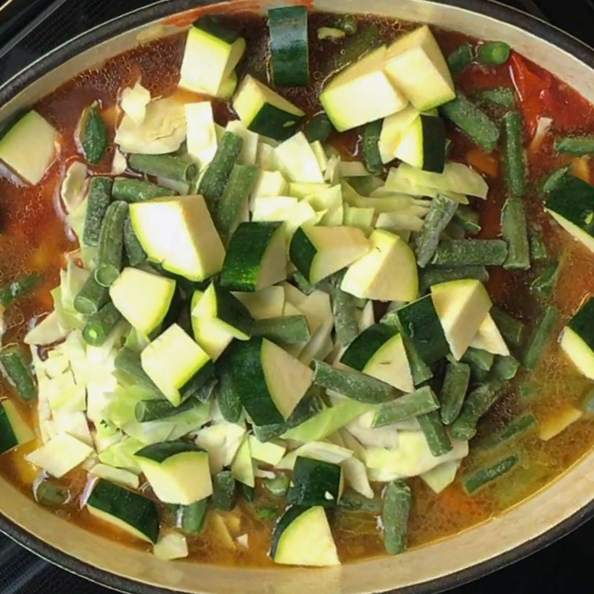 Soup simmering in a dutch, filled with chopped zucchini, green beans, and cabbage on a rich tomato-based broth.