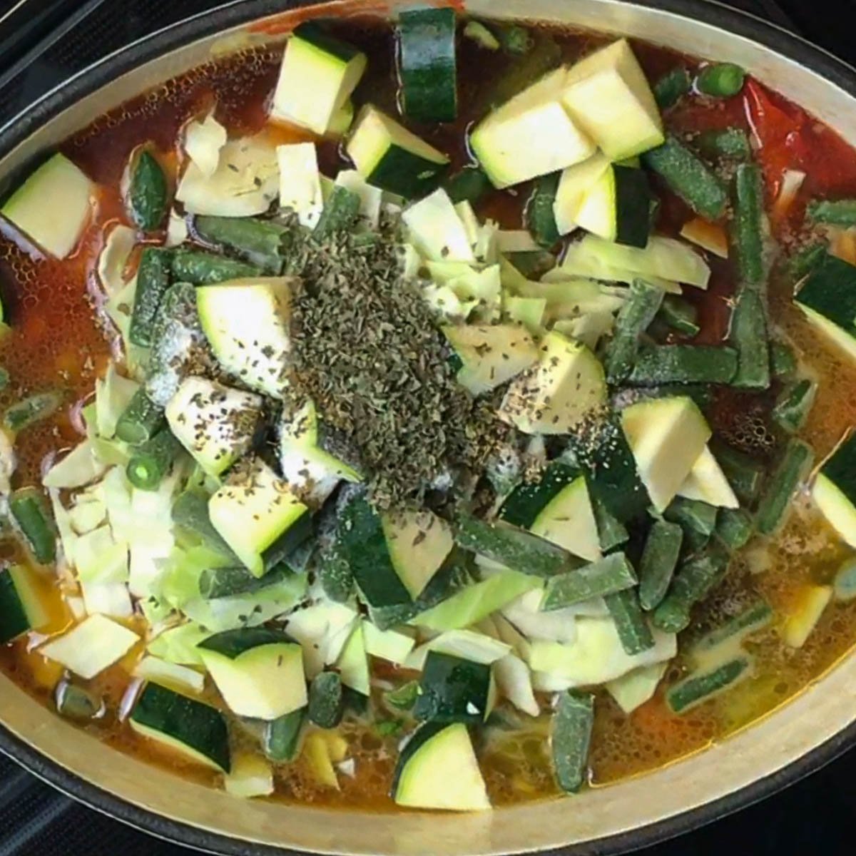 A pot of soup filled with chopped zucchini, green beans, and cabbage in a rich, red broth. A sprinkle of dried herbs and ground pepper are placed in the centre.