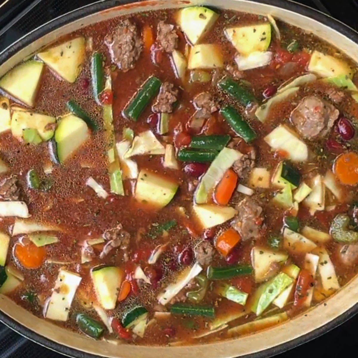 A hearty vegetable and meat soup simmers in a pot. Visible ingredients include chunks of zucchini, beans, carrots, and ground meat and sausage in a rich broth.