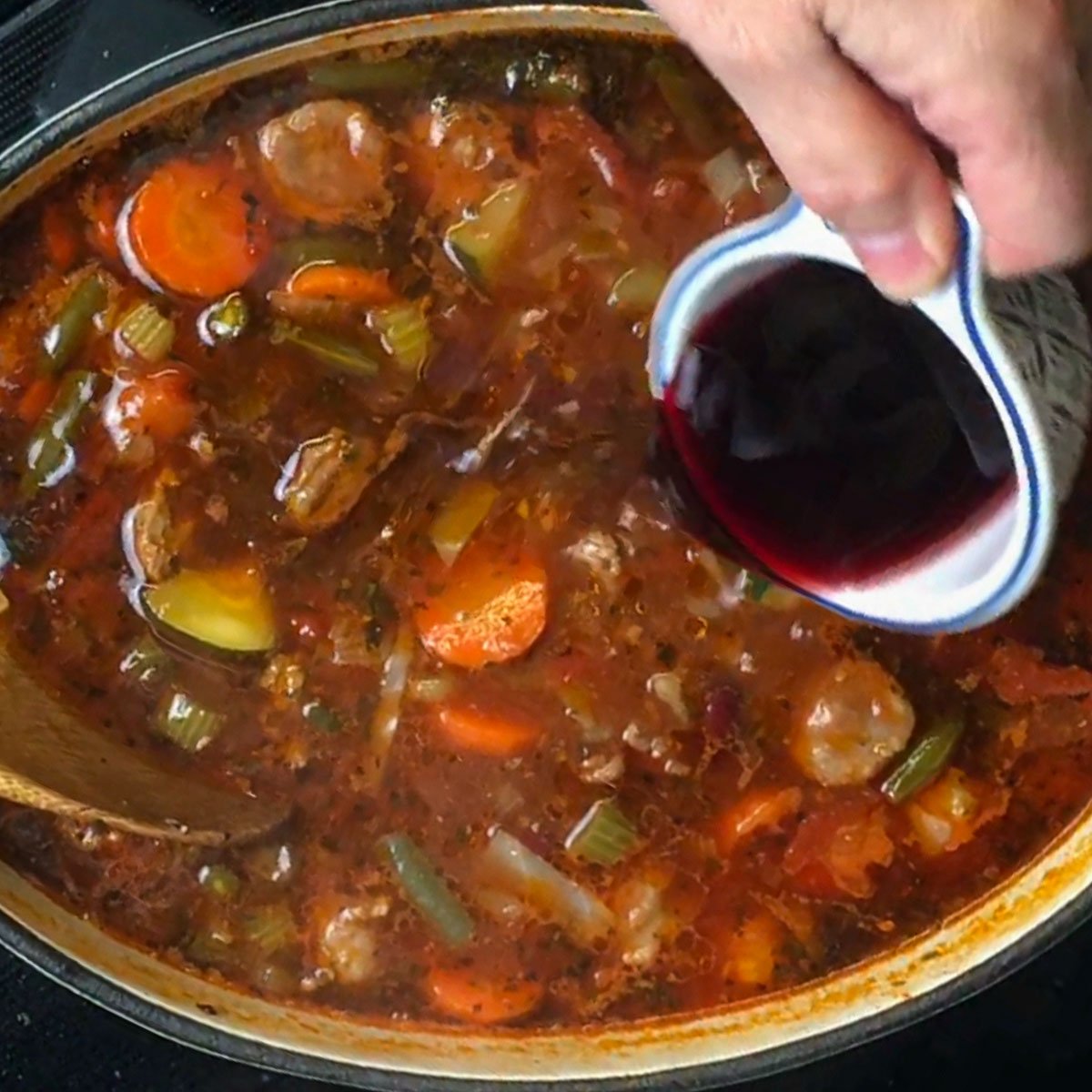 Close-up of a hand pouring red wine into a simmering pot of soup with vegetables, ground beef and sausage.