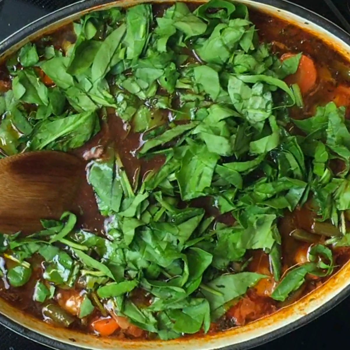 A pot of rich, tomato-based soup topped with fresh spinach. A wooden spoon stirs the mix.