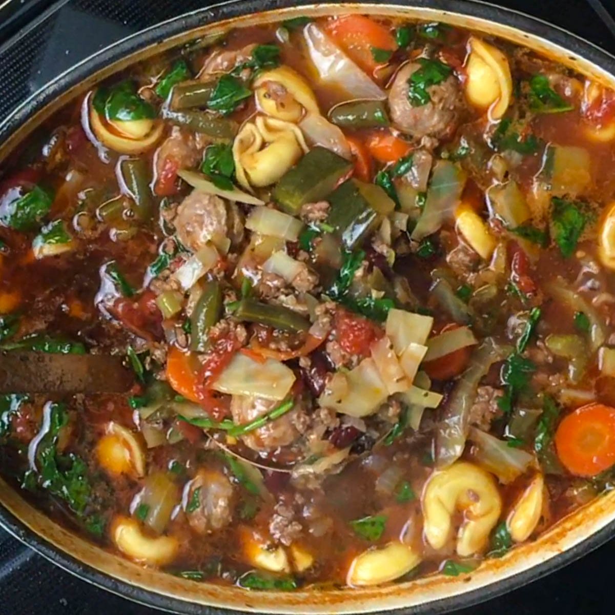 A dutch oven of soup with tortellini, ground beef, sausage, sliced vegetables, and herbs in a rich broth.