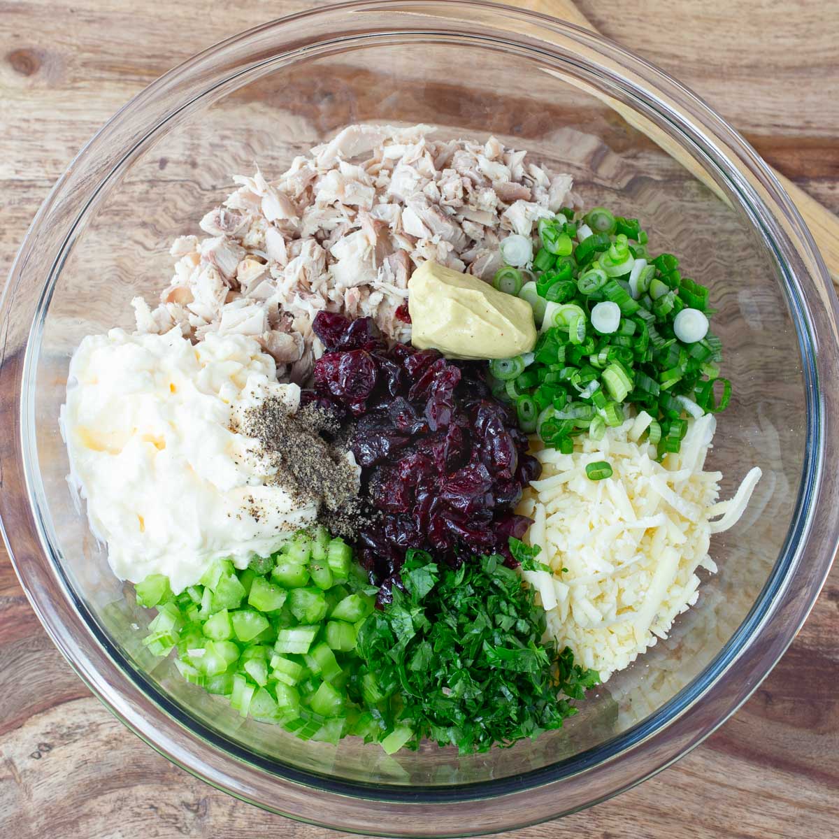 Glass bowl on wooden table with turkey salad ingredients: chopped chicken, mayonnaise, mustard, cranberries, celery, green onions, cilantro, cheese, and parsley.