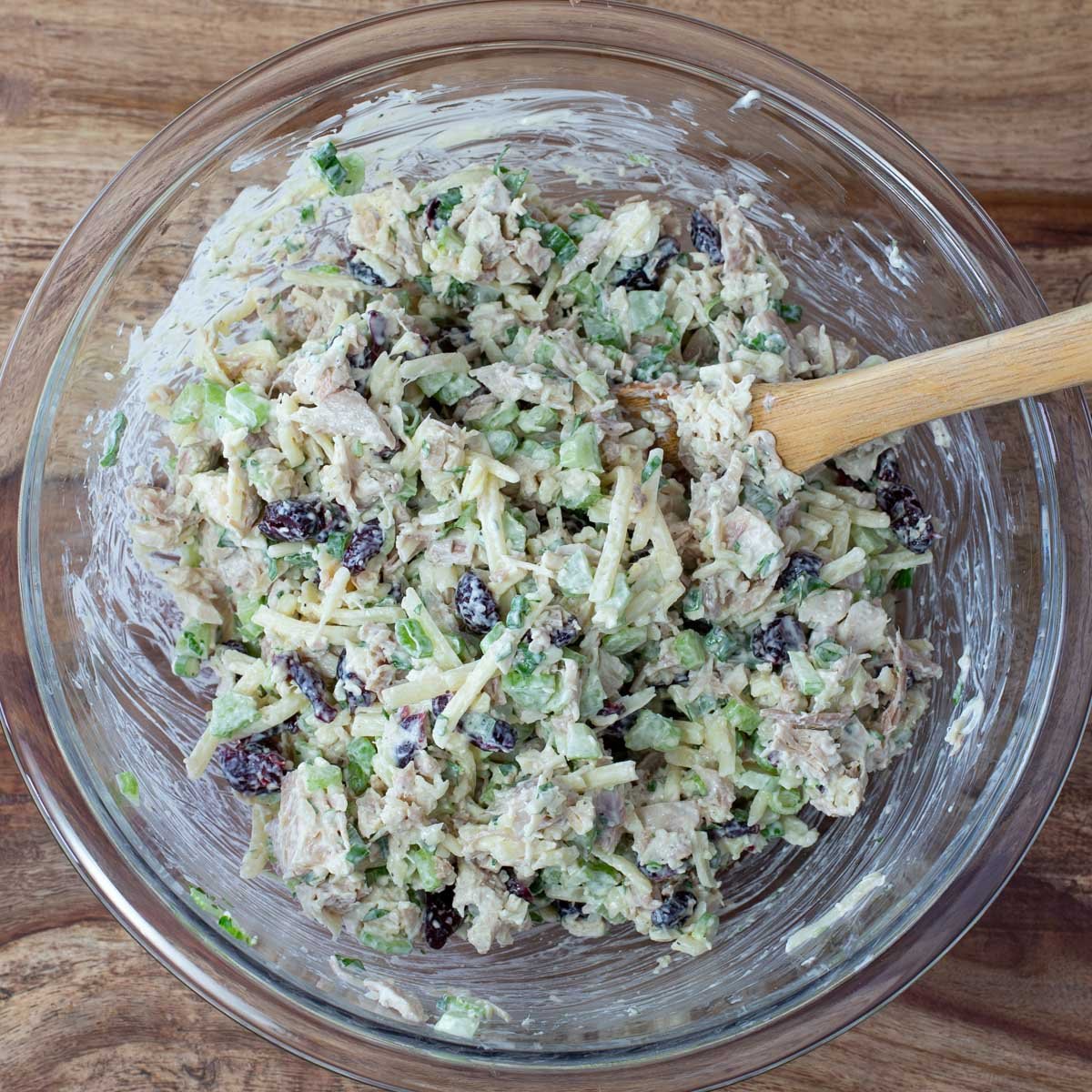 A glass bowl filled with creamy turkey salad, mixed with chopped celery, cranberries and shredded cheese, with a wooden spoon.