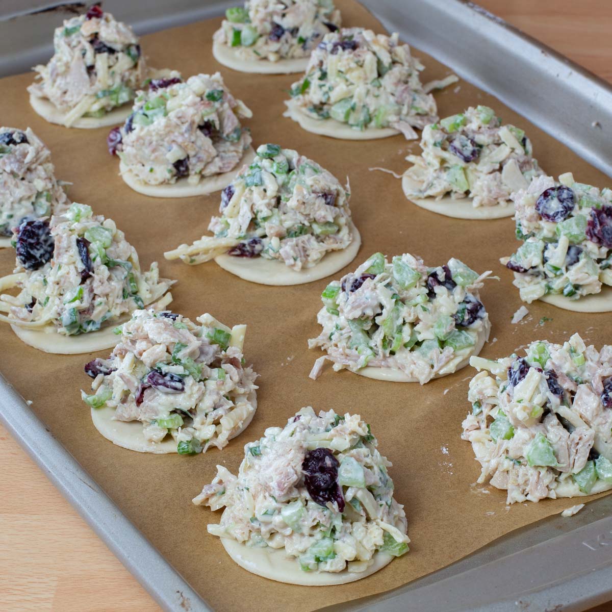 Rows of small, round pastries topped with a mixture of chopped turkey, cheese, celery, green onions, and cranberries on brown parchment paper lined baking sheet.