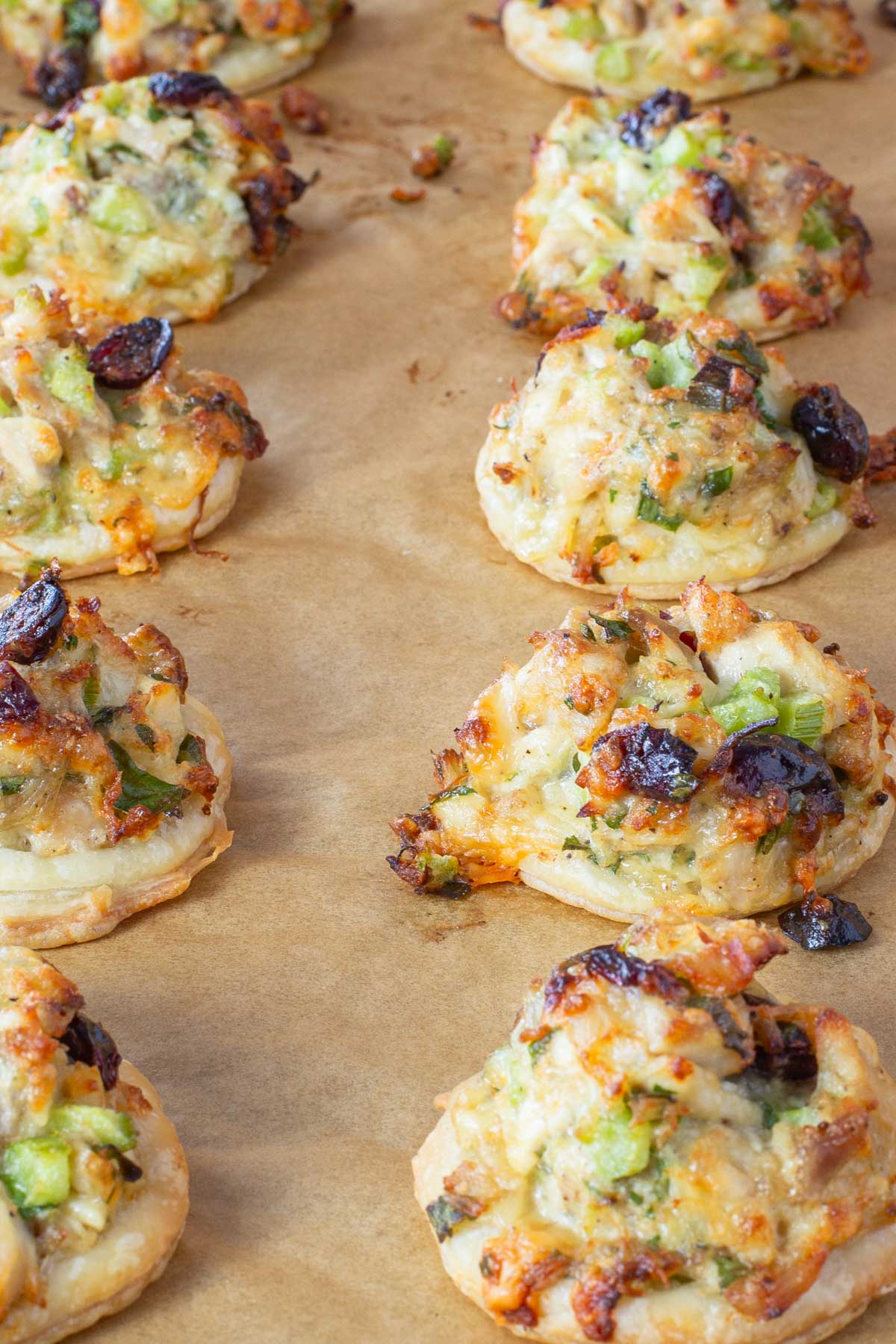 Rows of small, round pastries topped with chopped turkey, melted cheese, celery, green onions, and cranberries on brown parchment paper.