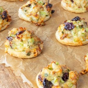 Rows of small, round pastries topped with chopped turkey, melted cheese, celery, green onions, and cranberries on brown parchment paper.