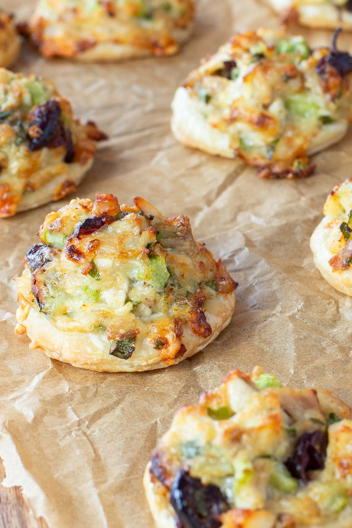 Rows of small, round pastries topped with chopped turkey, melted cheese, celery, green onions, and cranberries on brown parchment paper.