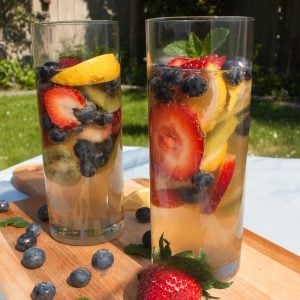 Two tall glasses filled with a refreshing fruit-infused wine containing strawberries, blueberries, lemon slices, and kiwi, set on a wooden board outdoors.