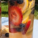 Tall glasses filled with a refreshing fruit-infused wine containing strawberries, blueberries, lemon slices, and kiwi, set on a wooden board.
