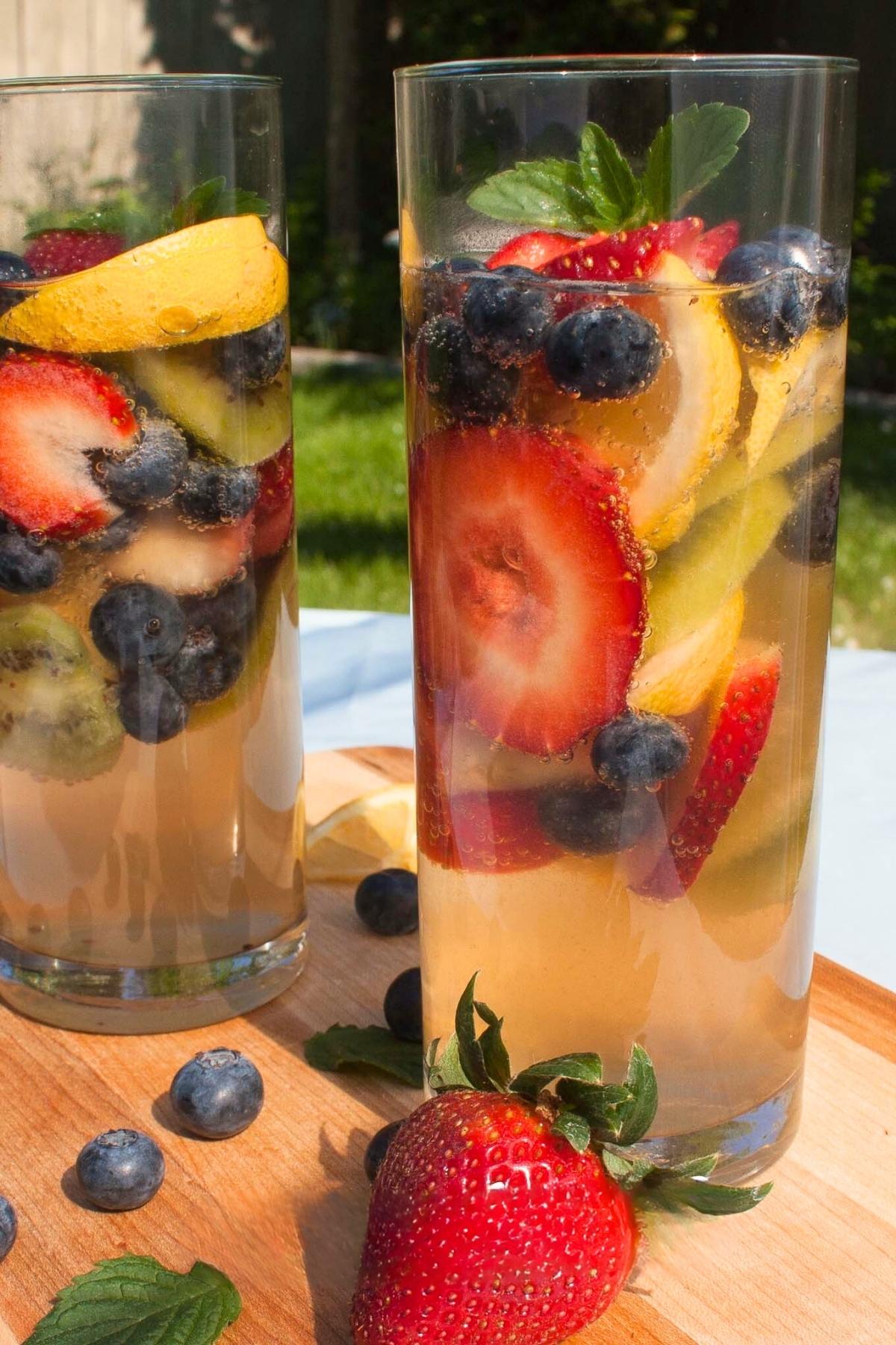 Two tall glasses filled with a refreshing fruit-infused wine containing strawberries, blueberries, lemon slices, and kiwi, set on a wooden board outdoors.