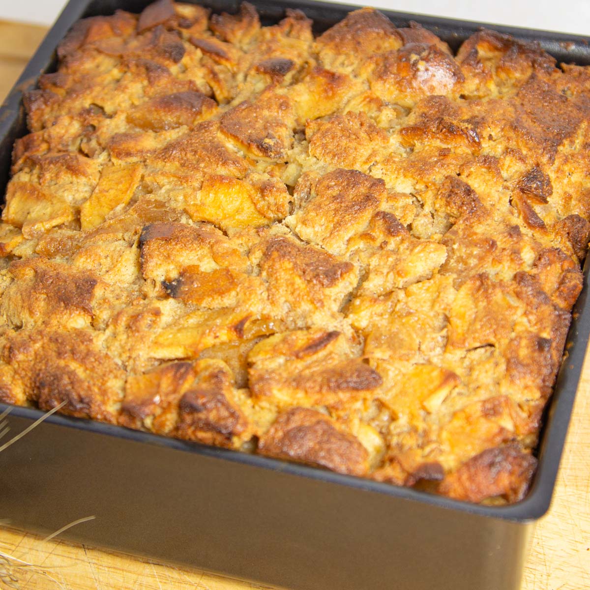 Golden brown bread pudding fills a square black baking dish. The top is crispy with visible chunks of bread and hints of caramelization.