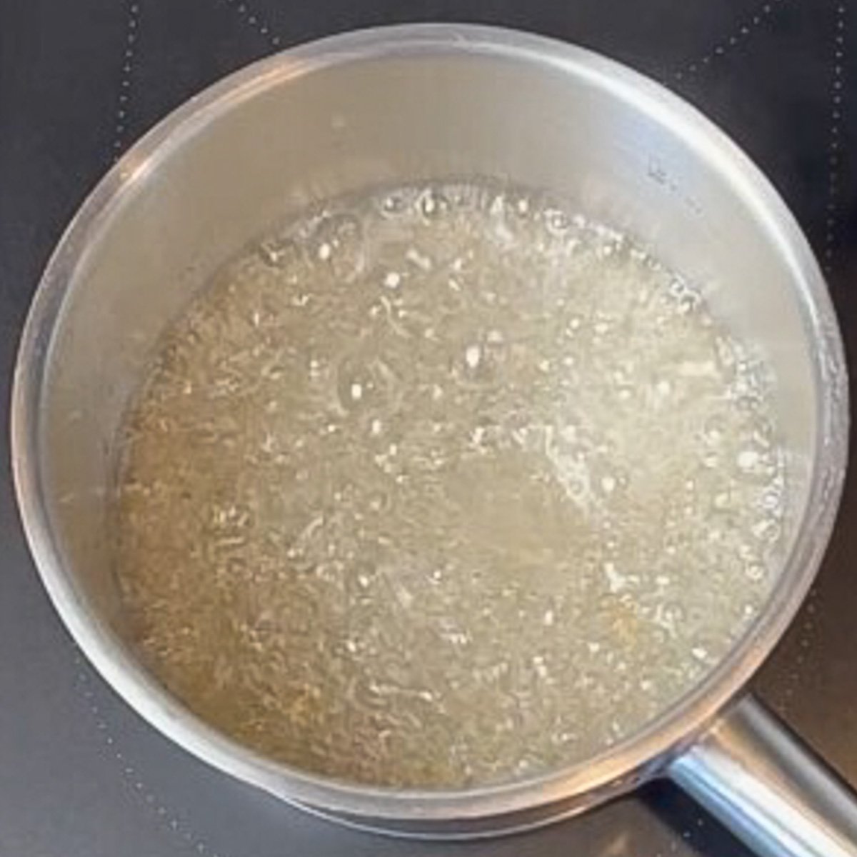 A steel saucepan filled with boiling mixture of sugar and water sits on a black stovetop.