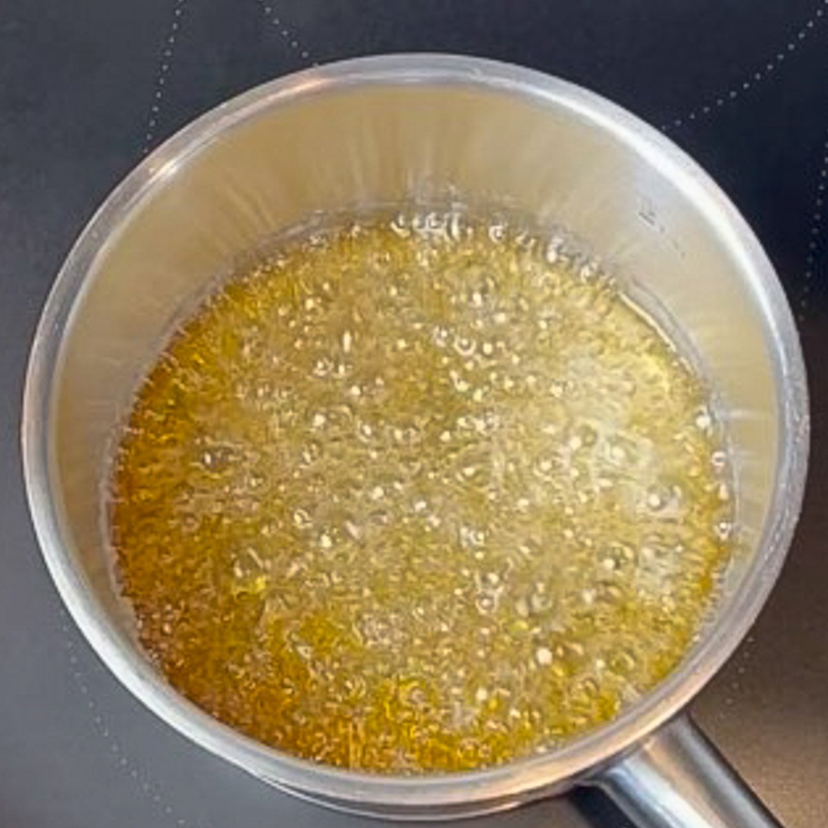 A steel saucepan filled with boiling mixture of amber colored sugar and water sits on a black stovetop.