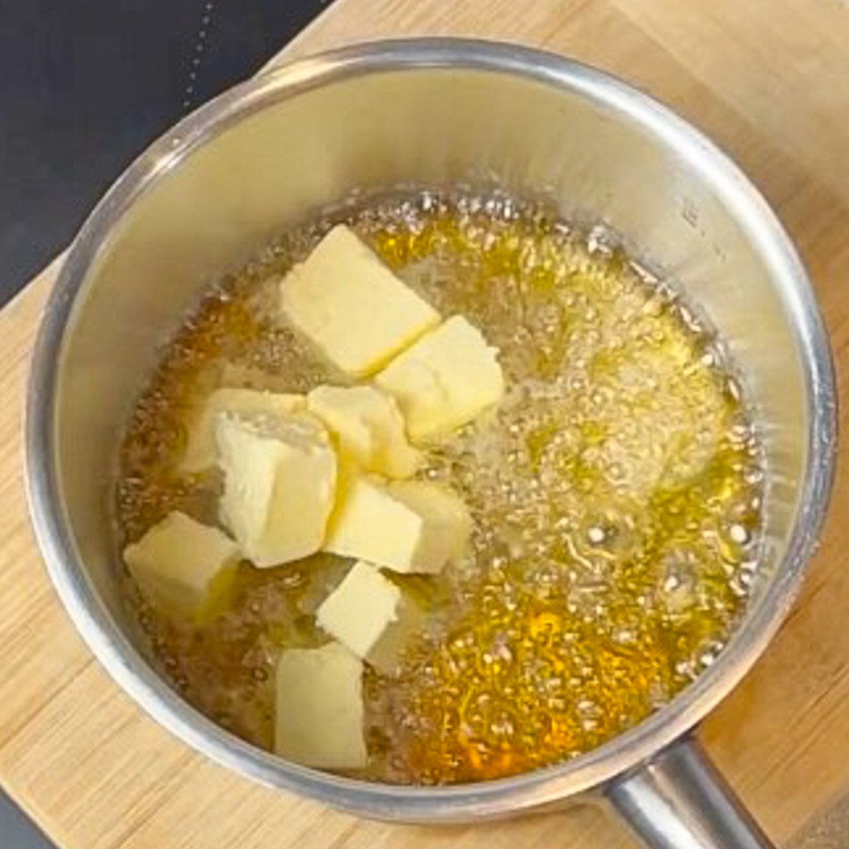A steel saucepan filled with boiling mixture of sugar and water sits on a wood board. Cubes of butter are added.