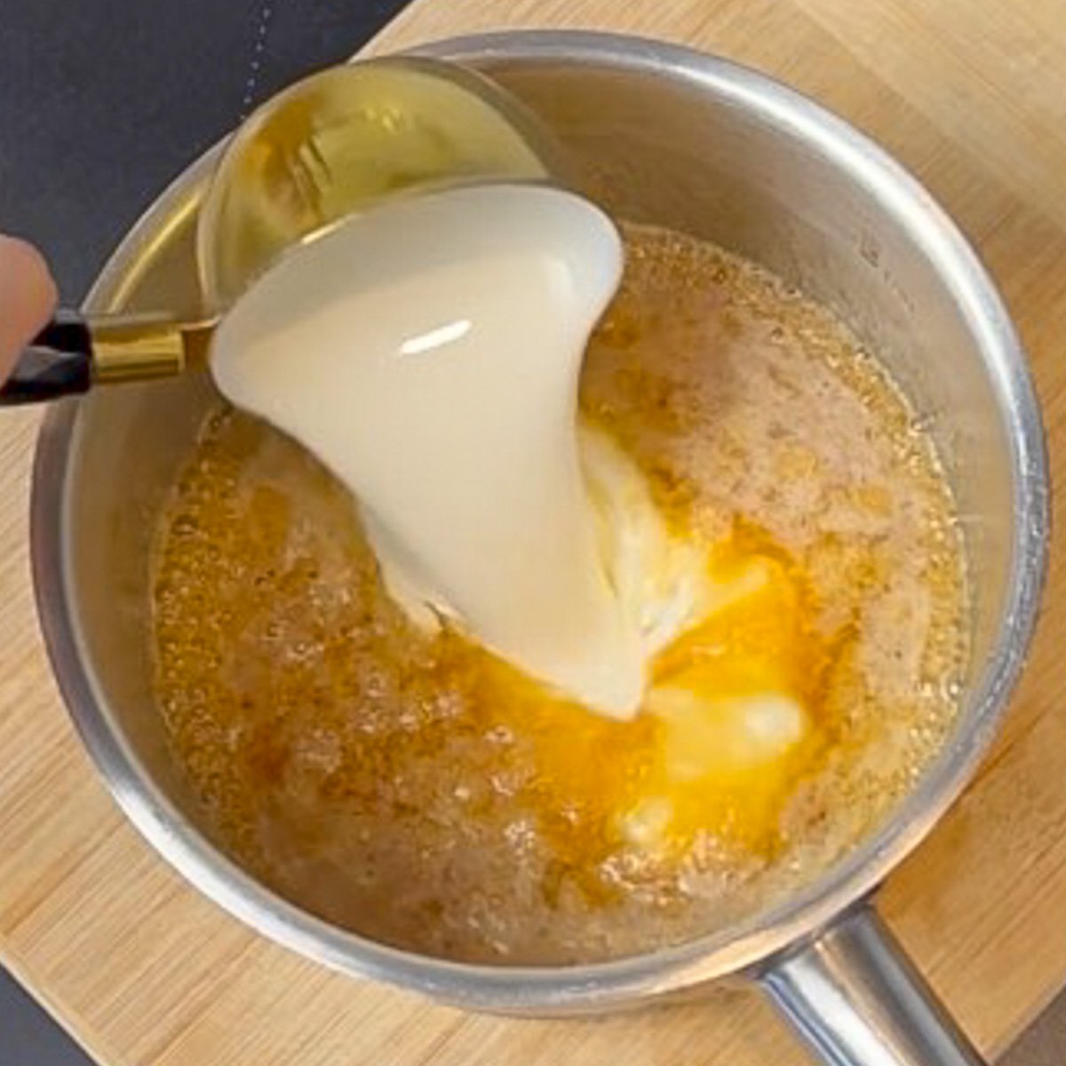 Cream being poured into a steel saucepan of bubbling caramel sauce.