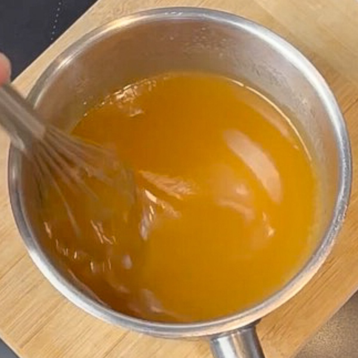 A steel saucepan filled with caramel sauce sits on a wood board. Whisk is stirring the mixture.
