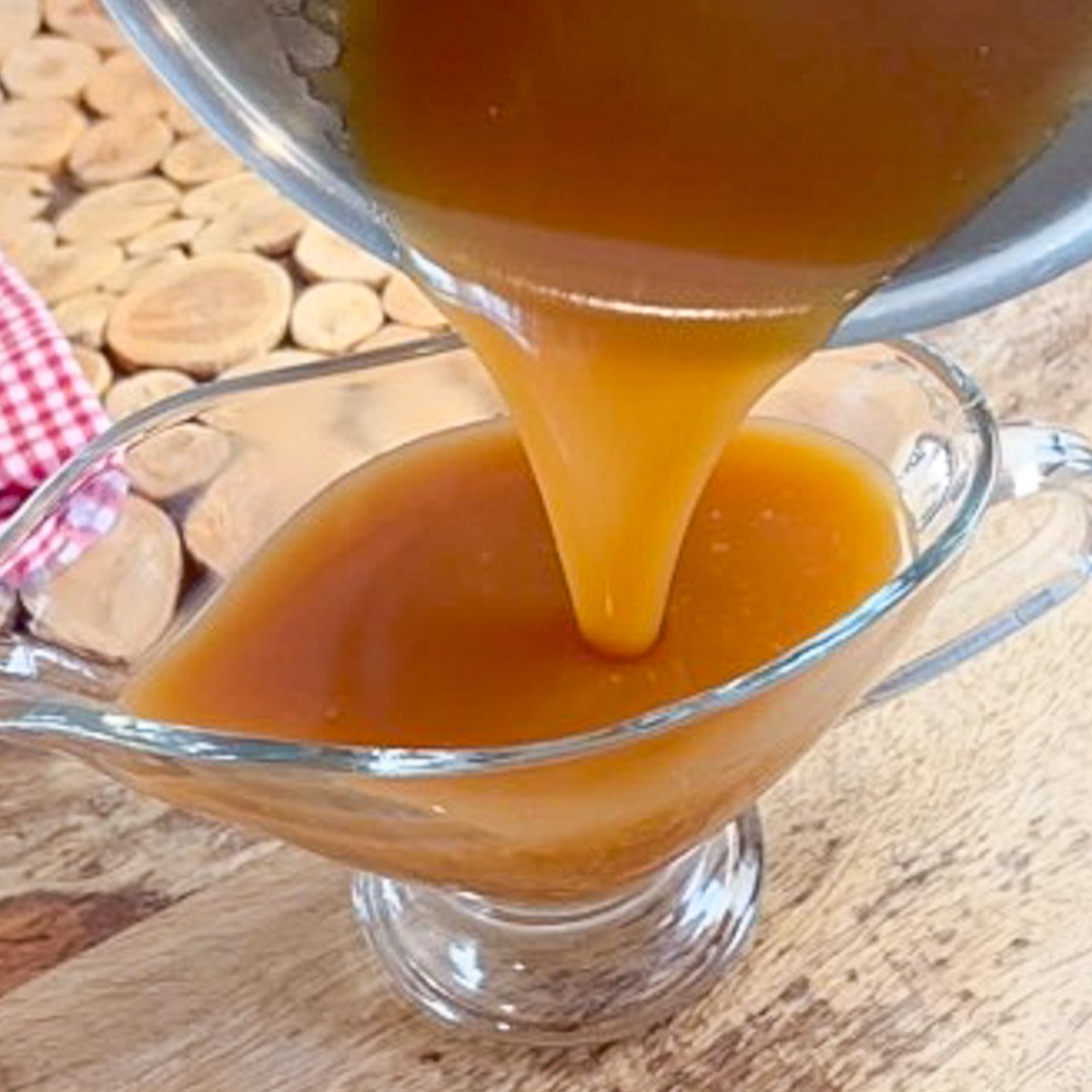 Caramel sauce is being poured from a pot into a glass serving dish on a wooden table.