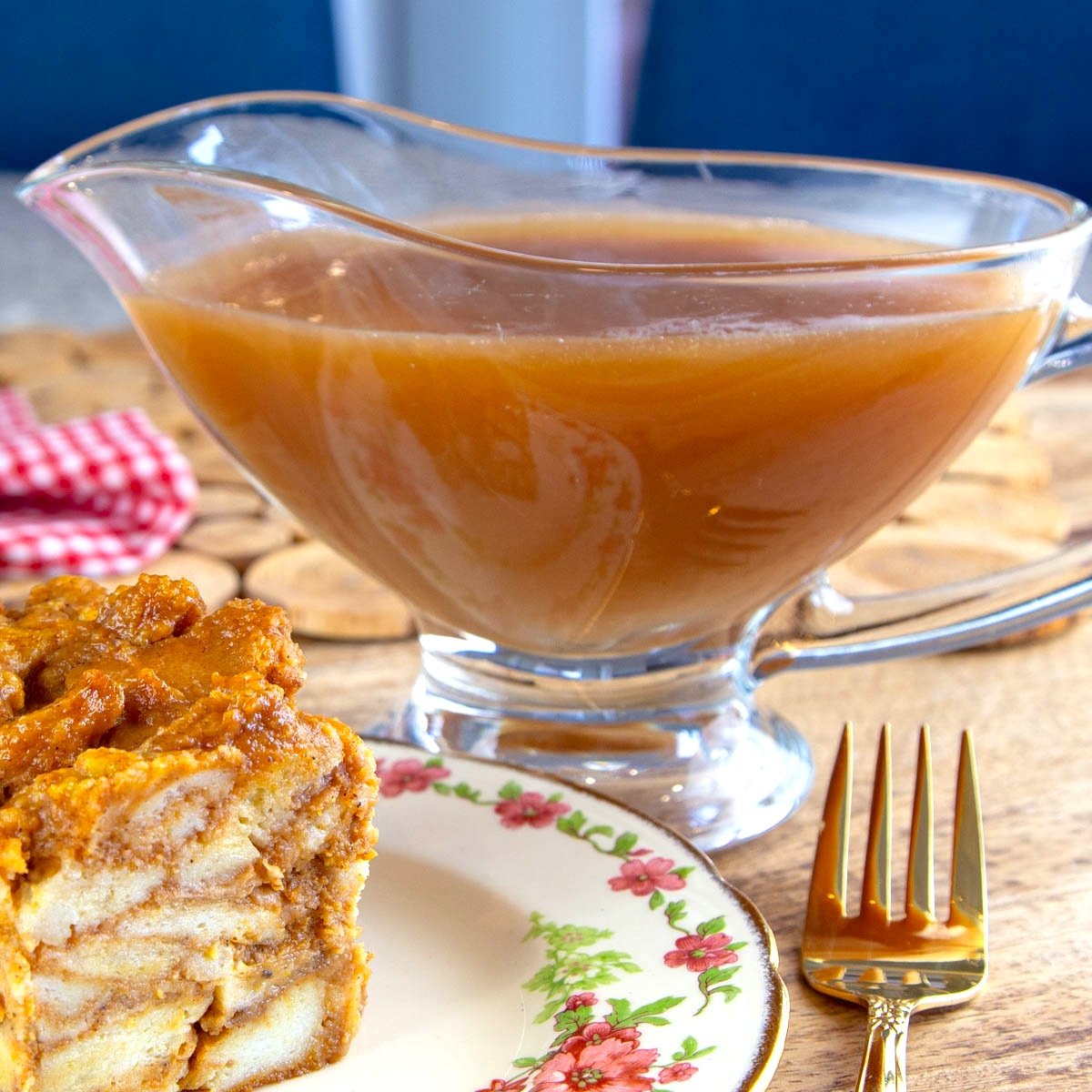 Glass serving dish filled with caramel sauce beside slice of bread pudding on a decorative plate.