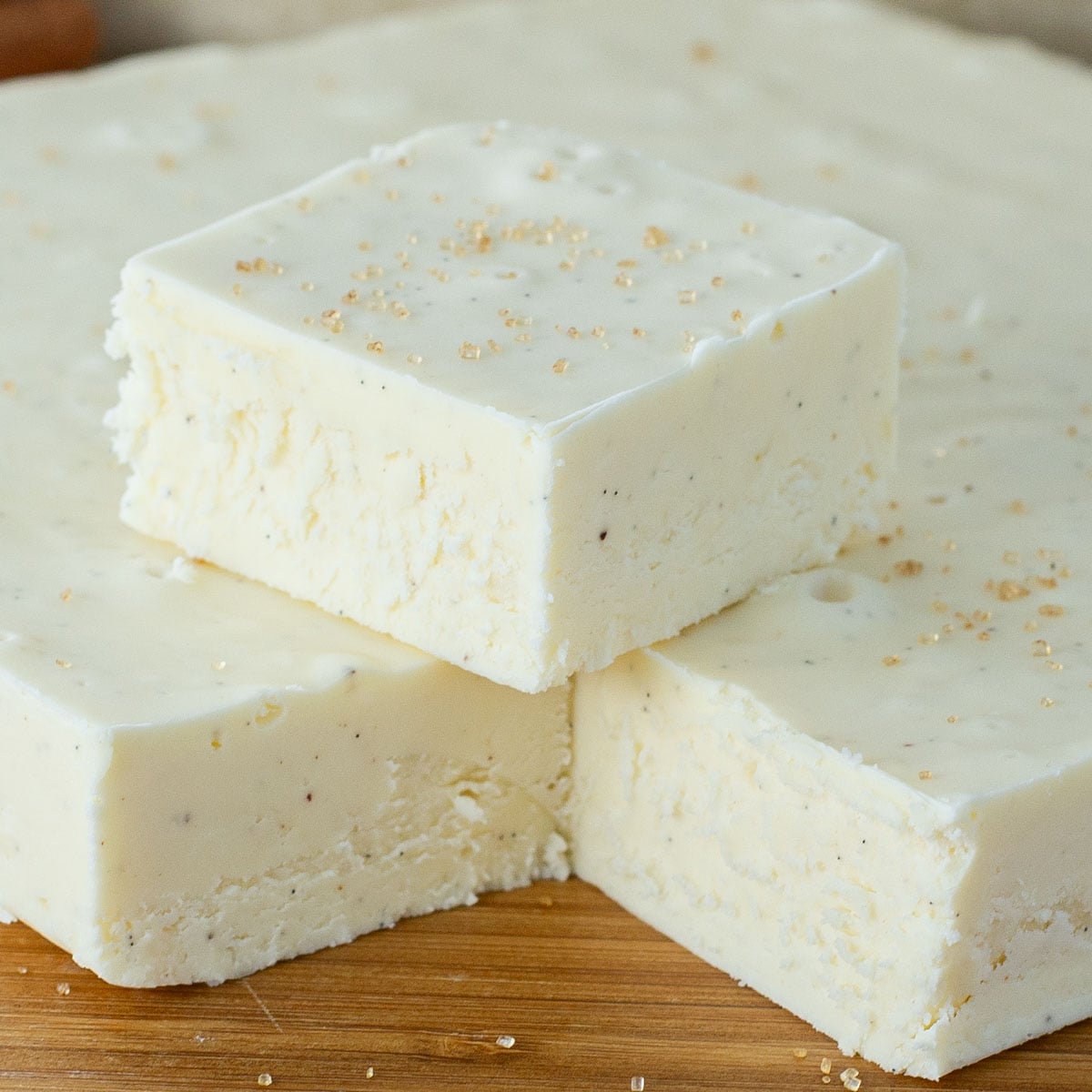 Squares of creamy white eggnog fudge are stacked full slab of fudge on a wooden board. The fudge is sprinkled with small golden sugar crystals.