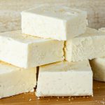 Stack of creamy white eggnog fudge squares topped with sugar crystals, placed on a wooden surface.