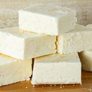 Stack of creamy white eggnog fudge squares topped with sugar crystals, placed on a wooden surface.