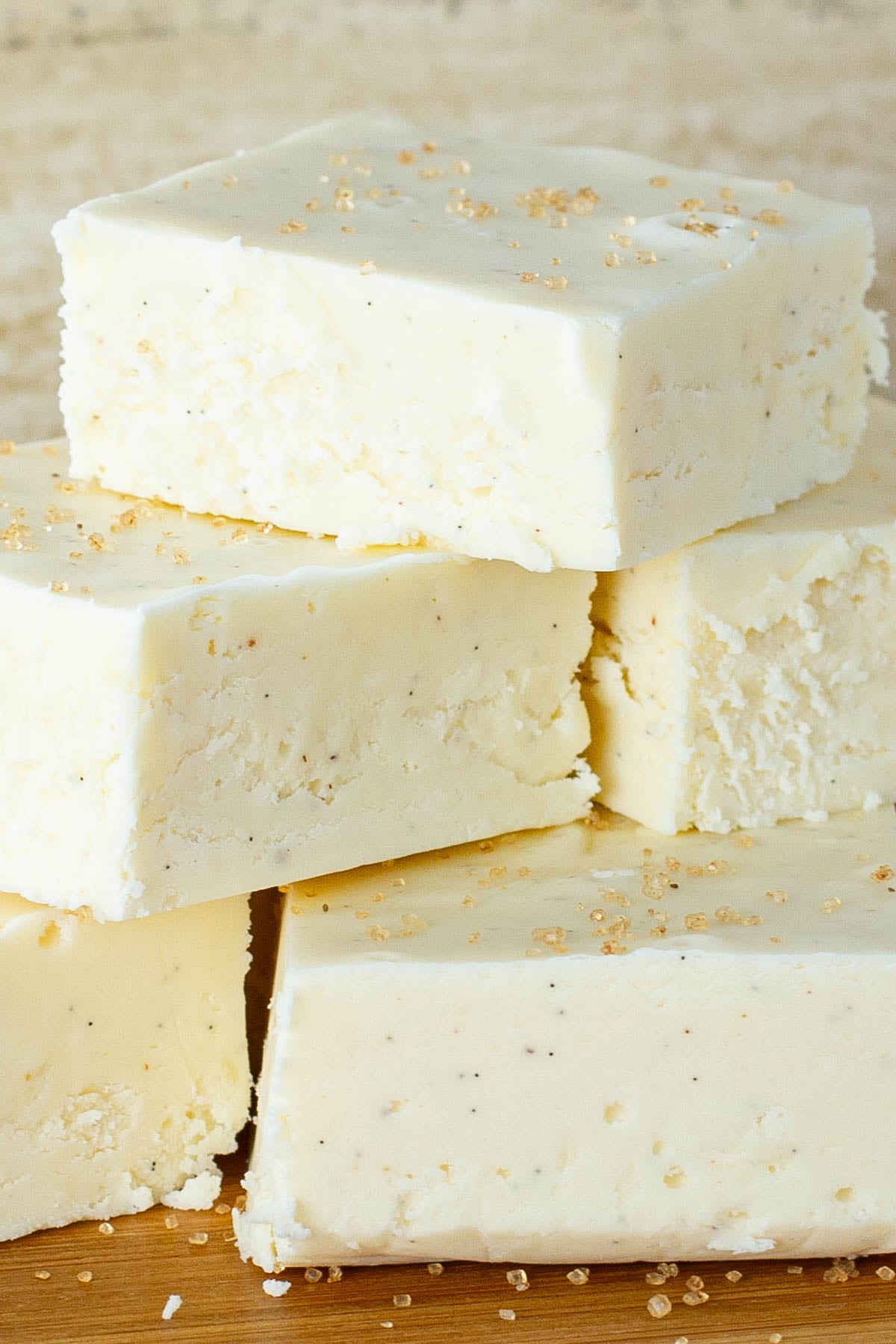 Stack of creamy white eggnog fudge squares topped with sugar crystals, placed on a wooden surface.