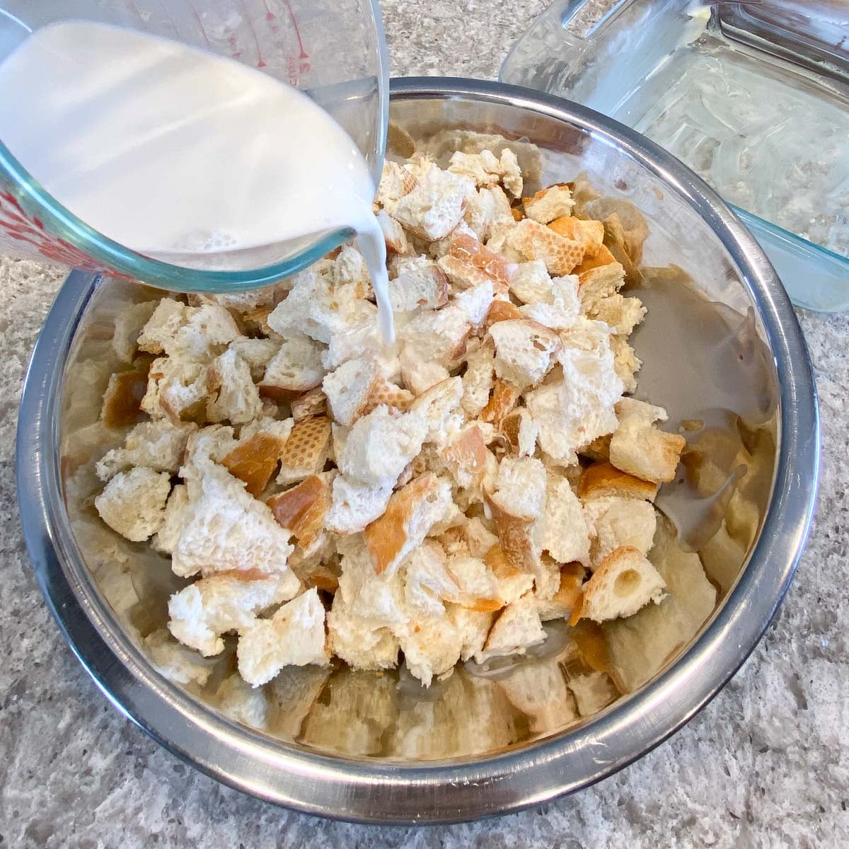 A glass measuring cup pours milk over torn bread pieces in a large metal bowl on a granite countertop.
