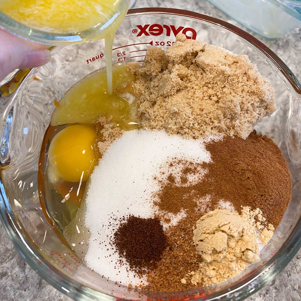 A glass bowl filled with ingredients for baking, including pumpkin puree, two eggs, brown sugar, white sugar, cinnamon, and ginger. Melted butter is being poured in.