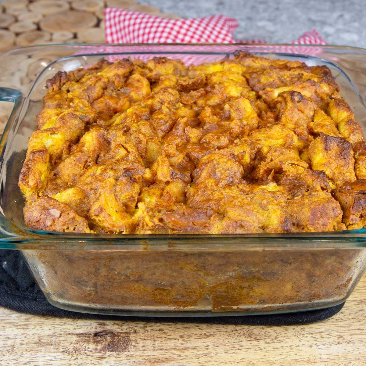 Golden pumpkin bread pudding in a clear glass dish on a wooden table, with a red checkered cloth in the background.