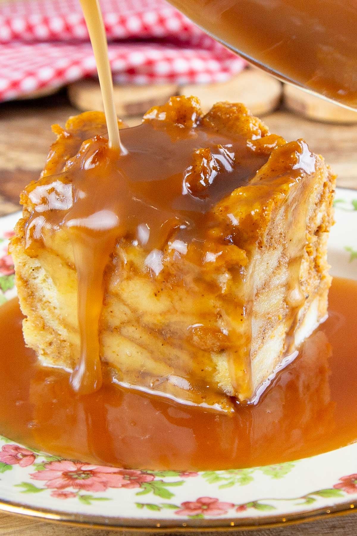 Rich caramel sauce being poured over a square piece of bread pudding on a floral plate.