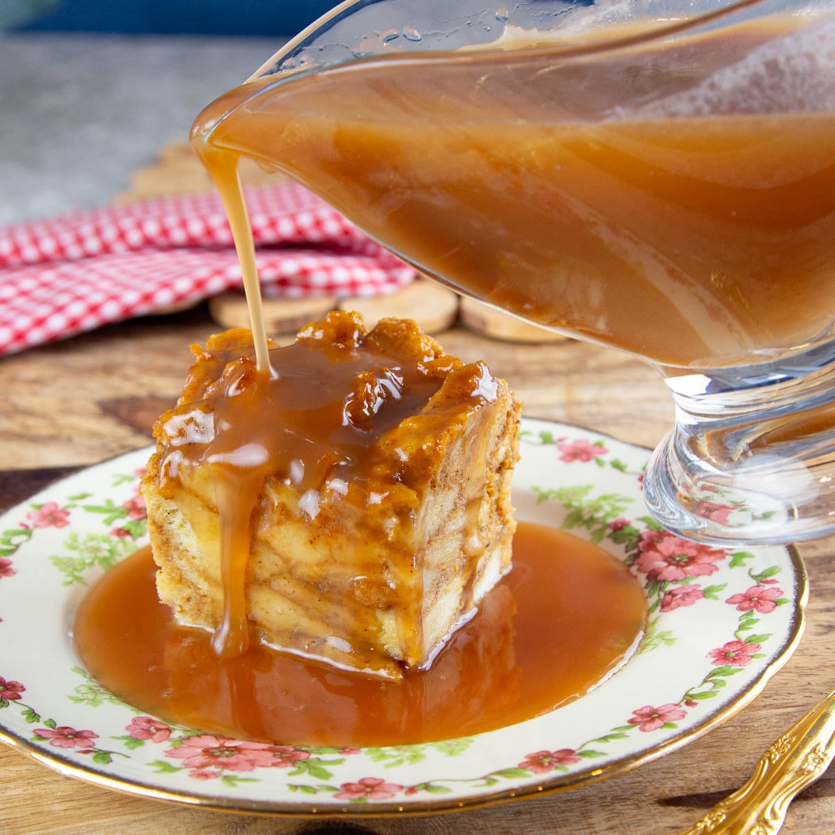 Rich caramel sauce being poured over a square piece of bread pudding on a floral plate.
