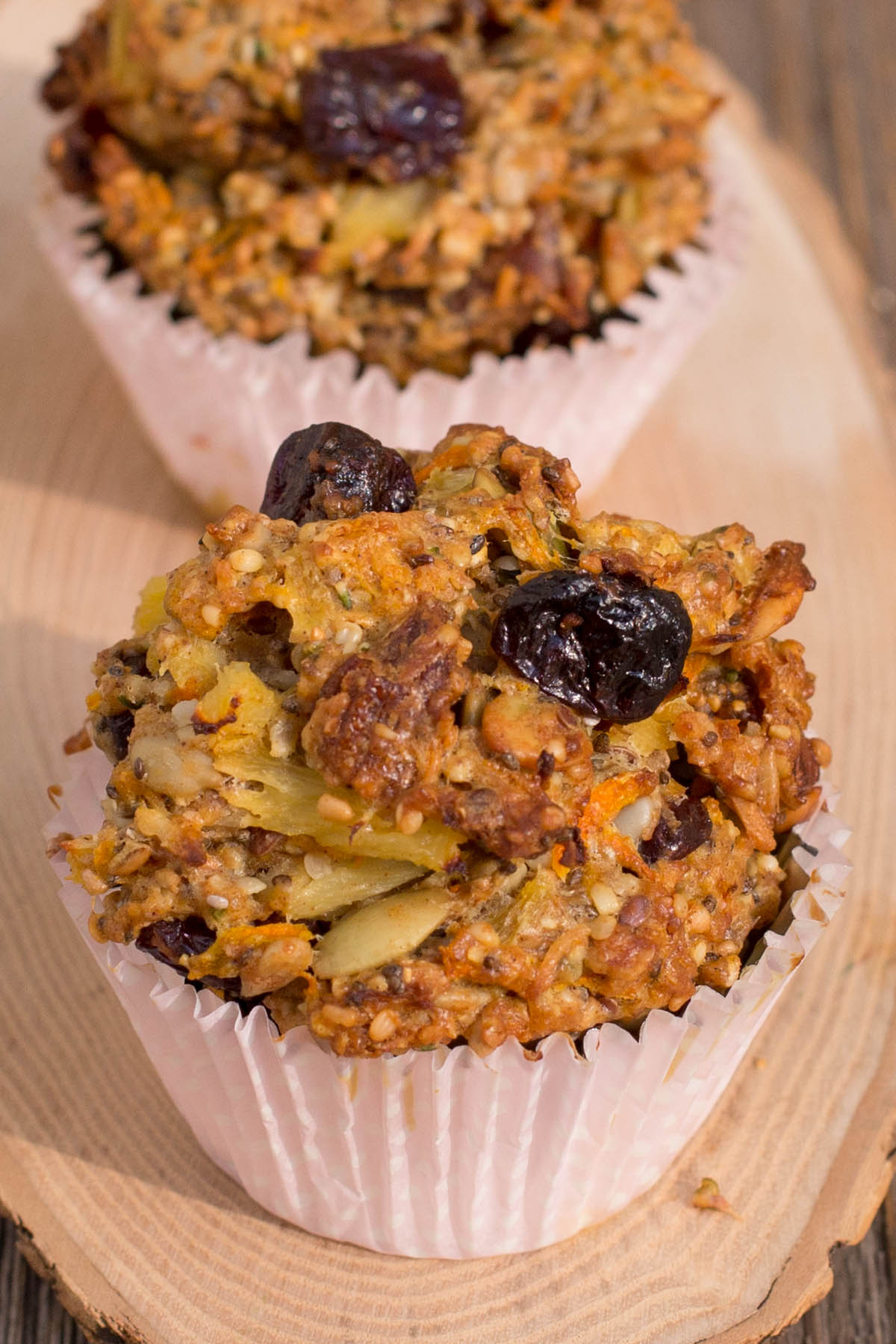 Hearty muffins with dried cherries, nuts, and seeds in paper liners on a wood board.
