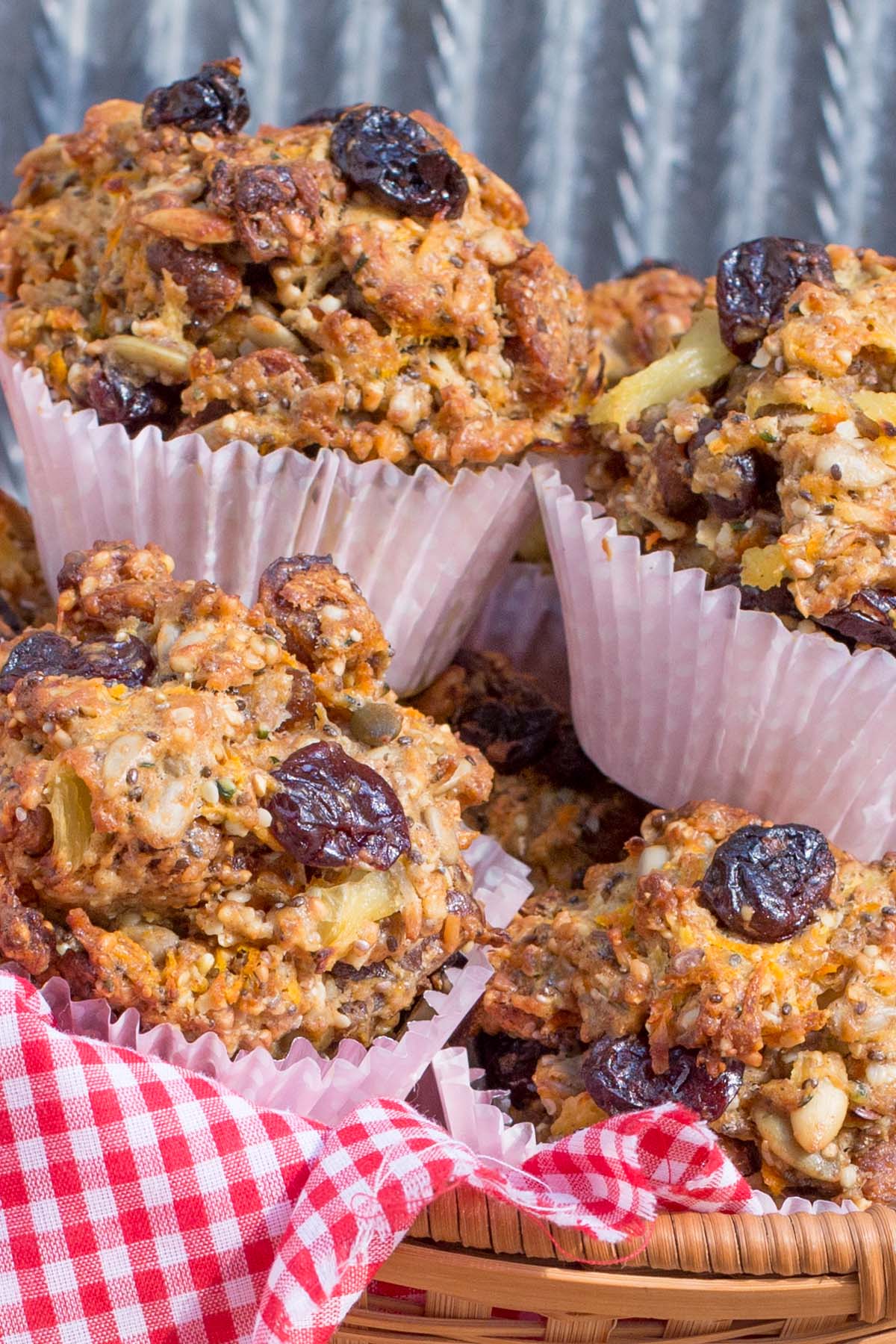 Hearty muffins with dried cherries, nuts, and seeds in paper liners fill a red gingham cloth lined basket.