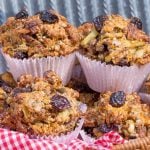 Hearty muffins with dried cherries, nuts, and seeds in paper liners fill a red gingham cloth lined basket.