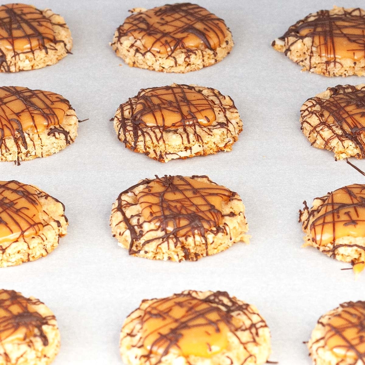 Oatmeal cookies topped with caramel and drizzled with chocolate rest on a parchment-lined baking sheet.