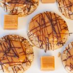 Oatmeal cookies topped with caramel and drizzled with chocolate on a white plate. Caramel squares are placed between the cookies.