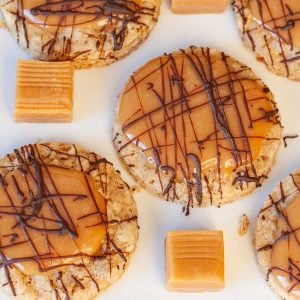 Oatmeal cookies topped with caramel and drizzled with chocolate on a white plate. Caramel squares are placed between the cookies.