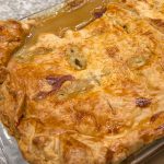 Golden brown pastry covers a homemade chicken pie in a rectangular dish, with gravy oozing out.